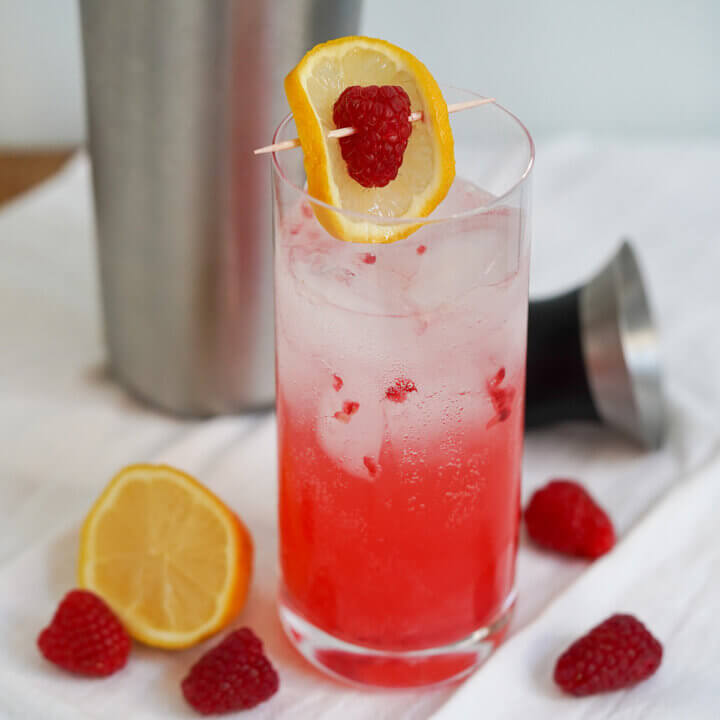 glass of raspberry collins cocktail with raspberries and cut lemon to side of glass.