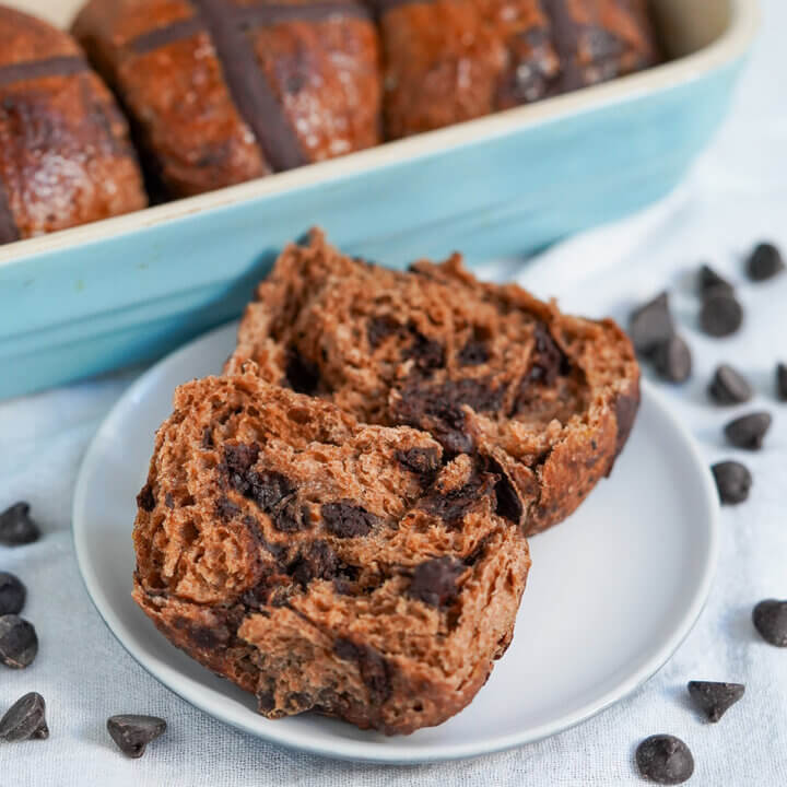 chocolate hot cross bun on plate with chocolate chips around and part view of more in dish behind.